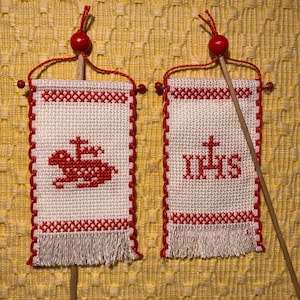 Könnte beinhalten: Zwei weiße Kreuzstichbanner mit roten Akzenten und Fransen. Ein Banner zeigt ein rotes Lamm mit einem Kreuz, das andere zeigt die Buchstaben IHS in Rot. Jedes Banner ist an einem Holzstab mit einer roten Perle befestigt.