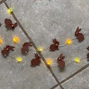 Könnte beinhalten: Eine Lichterkette mit braunen Hasenformen und gelben und grünen Glühbirnen. Die Hasen haben rote Schleifen und goldene Glöckchen.