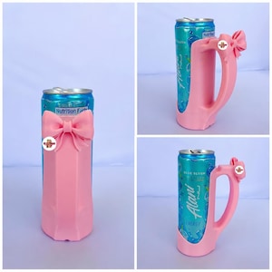 May include: A pink can holder with a bow design, holding a blue and teal can of Alani Nu Blue Slush energy drink. The can holder has a handle and a bow accent. The can has the words "Blue Slush" and "Energy" on it.