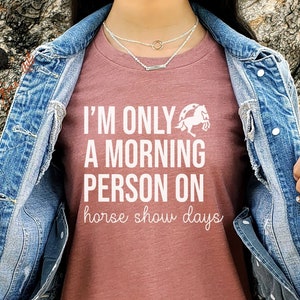 K&ouml;nnte beinhalten: Ein T-Shirt in Heide-Mauve mit dem Text "I'm only a morning person on horse show days" und einer wei&szlig;en Silhouette eines springenden Pferdes &uuml;ber einem Hufeisen.