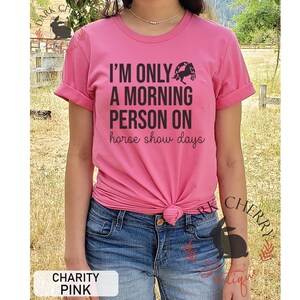 K&ouml;nnte beinhalten: Pinkes T-Shirt mit einem schwarzen Aufdruck, auf dem "Ich bin nur ein Morgenmensch an Pferdeshow-Tagen" steht. Der Aufdruck enth&auml;lt auch eine Silhouette eines Pferdes, das &uuml;ber ein Hufeisen springt.