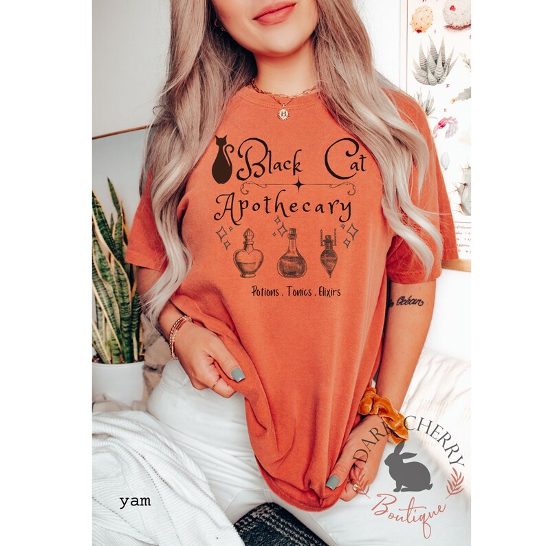 Puede incluir: Una camiseta naranja quemada con un gr&aacute;fico de gato negro y el texto "Black Cat Apothecary" con tres frascos de poci&oacute;n y el texto "Potions. Tonics. Elixirs".