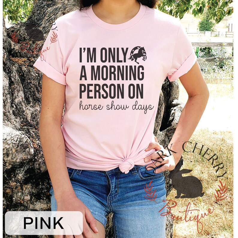 K&ouml;nnte beinhalten: Pinkes T-Shirt mit einem schwarzen Aufdruck, der "Ich bin nur ein Morgenmensch an Pferdeshow-Tagen" sagt, mit einer Silhouette eines Pferdes, das &uuml;ber eine H&uuml;rde springt.