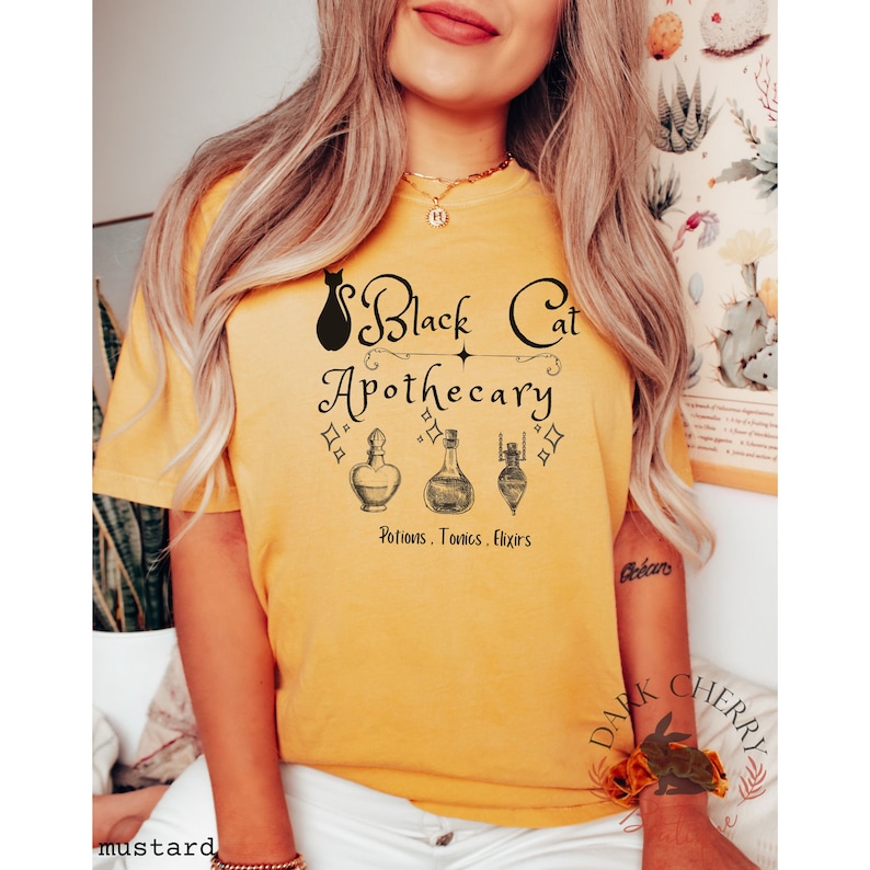 Puede incluir: Una camiseta amarilla mostaza con un gr&aacute;fico de un gato negro y el texto "Black Cat Apothecary" con una estrella y tres frascos de pociones. El texto debajo de los frascos dice "Potions, Tonics, Elixirs".