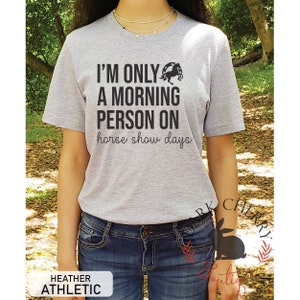 K&ouml;nnte beinhalten: Graues T-Shirt mit dem Text "Ich bin nur ein Morgenmensch an Pferdeshow-Tagen" und einer Silhouette eines Pferdes, das &uuml;ber eine H&uuml;rde springt.