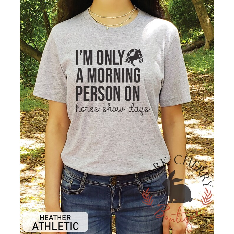 K&ouml;nnte beinhalten: Graues T-Shirt mit dem Text "Ich bin nur ein Morgenmensch an Pferdeshow-Tagen" und einer Silhouette eines Pferdes, das &uuml;ber eine H&uuml;rde springt.