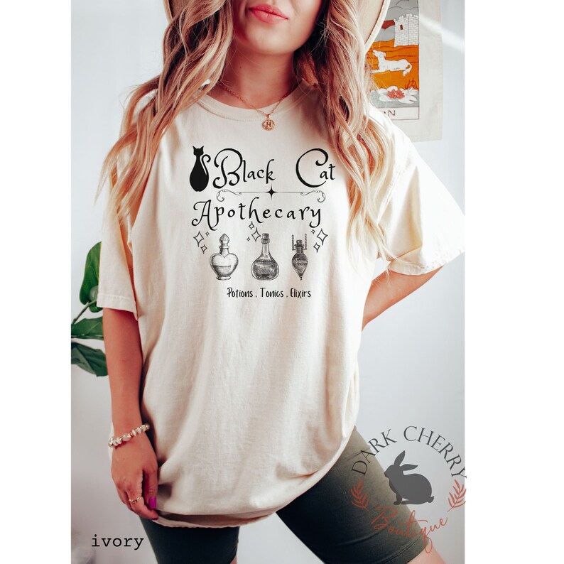 Puede incluir: Una camiseta blanca con un gato negro y el texto "Black Cat Apothecary" y "Potions. Tonics. Elixirs".