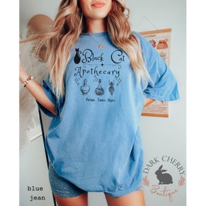 Puede incluir: Una camiseta azul con un gr&aacute;fico de un gato negro y el texto "Black Cat Apothecary" con una estrella y tres frascos de pociones. El texto debajo de los frascos dice "Potions. Tonics. Elixirs".