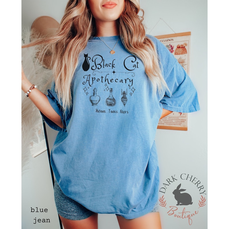 Puede incluir: Una camiseta azul con un gr&aacute;fico de un gato negro y el texto "Black Cat Apothecary" con una estrella y tres frascos de pociones. El texto debajo de los frascos dice "Potions. Tonics. Elixirs".