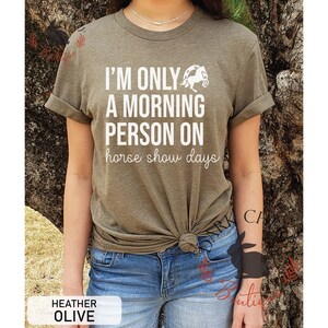 K&ouml;nnte beinhalten: Ein gr&uuml;nes T-Shirt mit dem Text "I'm only a morning person on horse show days" in Wei&szlig;. Neben dem Text befindet sich eine wei&szlig;e Silhouette eines Pferdes. Das Shirt ist in der Taille gebunden und das Model tr&auml;gt eine blaue Jeans.