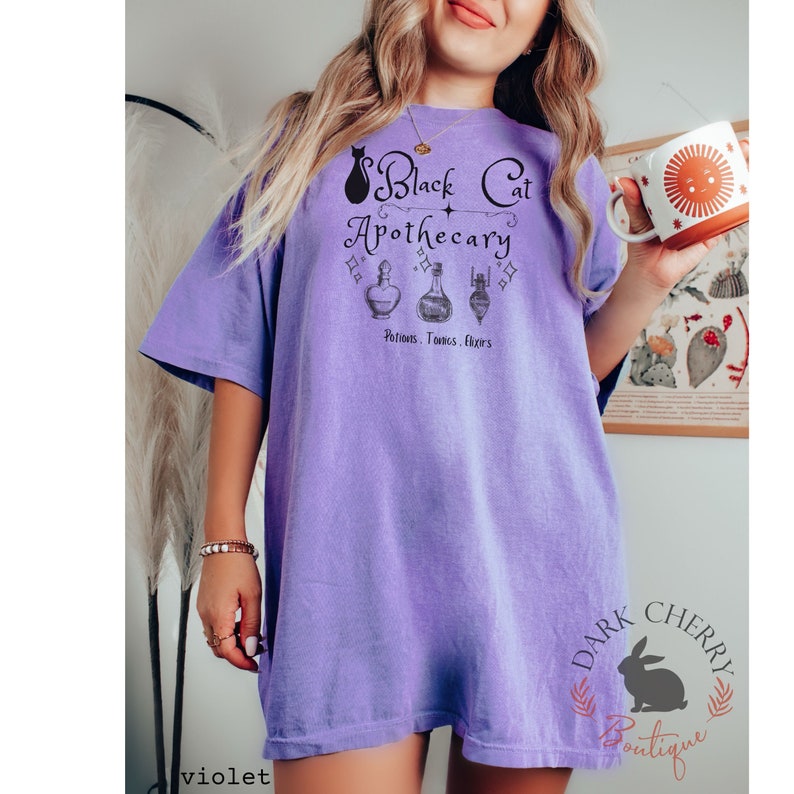Puede incluir: Camiseta morada de gran tama&ntilde;o con un gr&aacute;fico de un gato negro y el texto "Black Cat Apothecary" con tres frascos de pociones y el texto "Potions, Tonics, Elixirs". La camiseta tiene un aspecto desgastado y un corte holgado.