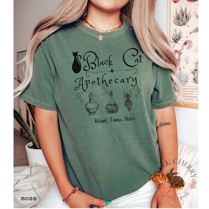 Puede incluir: Una camiseta verde con un gr&aacute;fico de un gato negro y el texto "Black Cat Apothecary Potions, Tonics, Elixirs".