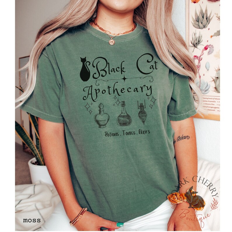 Puede incluir: Una camiseta verde con un gr&aacute;fico de un gato negro y el texto "Black Cat Apothecary Potions, Tonics, Elixirs".