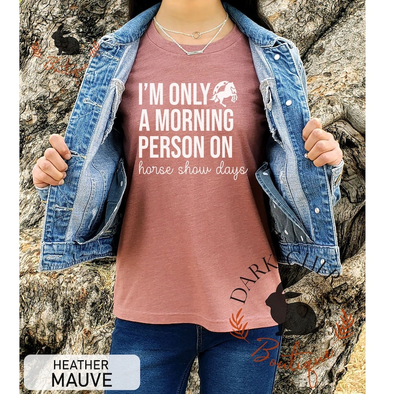 K&ouml;nnte beinhalten: Ein T-Shirt in Heide-Mauve mit dem Text "I'm only a morning person on horse show days" und einer wei&szlig;en Silhouette eines springenden Pferdes &uuml;ber einem Hufeisen.