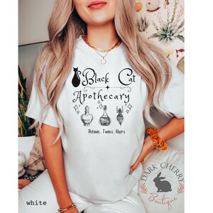 Puede incluir: Camiseta blanca con un gr&aacute;fico de gato negro y el texto "Black Cat Apothecary" con tres frascos de poci&oacute;n y el texto "Potions, Tonics, Elixirs".