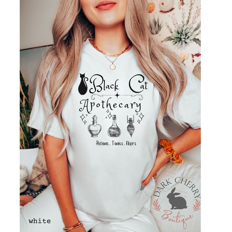 Puede incluir: Camiseta blanca con un gr&aacute;fico de gato negro y el texto "Black Cat Apothecary" con tres frascos de poci&oacute;n y el texto "Potions, Tonics, Elixirs".