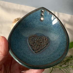 May include: A teal ceramic dish with a heart-shaped design in the centre. The dish has a teardrop shape with three small holes near the top. The rim is a darker brown colour.