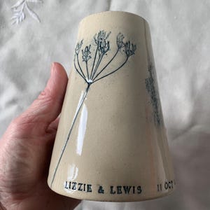 May include: A beige ceramic vase with a flared shape, featuring a blue floral design. The vase has the text "LIZZIE & LEWIS 11 OCT" printed on it. The vase is held by a person.
