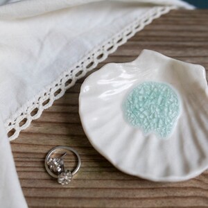 May include: White ceramic seashell dish with a light blue, crackled glaze in the center. The dish is on a wooden surface.