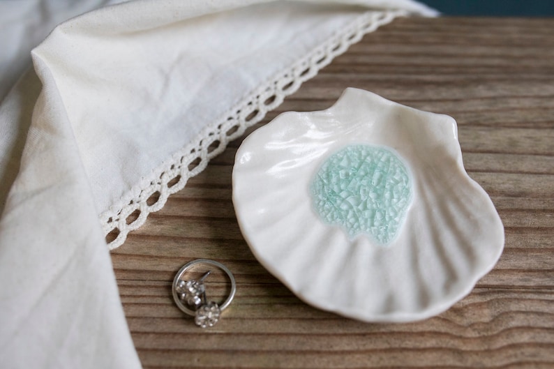 May include: White ceramic seashell dish with a light blue, crackled glaze in the center. The dish is on a wooden surface.