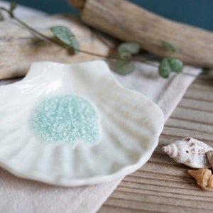 May include: A white ceramic dish shaped like a seashell with a light blue glaze in the center. The dish is sitting on a light brown surface with a seashell next to it.
