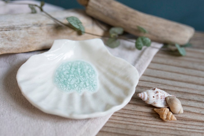 May include: A white ceramic dish shaped like a seashell with a light blue glaze in the center. The dish is sitting on a light brown surface with a seashell next to it.