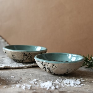 Puede incluir: Dos cuencos pequeños de cerámica blanca con un esmalte azul claro. Los cuencos tienen un acabado texturizado y craquelado y están sobre una superficie de madera.