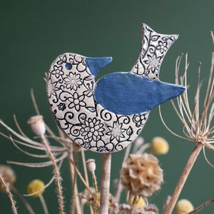 Könnte beinhalten: Eine Keramik-Vogelfigur mit blauem Körper und weißem Blumenmuster. Der Vogel sitzt auf einem Holzstab.