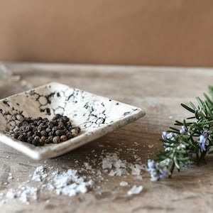 May include: A small white ceramic dish with black speckles, filled with black peppercorns. The dish is sitting on a wooden surface with white salt crystals scattered around it. A sprig of rosemary with purple flowers is in the background.
