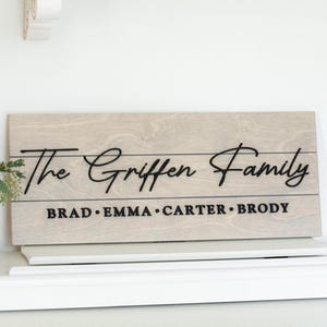 May include: A light wood sign with the text "The Griffen Family" in black cursive lettering. Below the family name, the names "Brad, Emma, Carter, Brody" are listed in black.