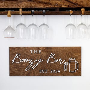 May include: A rustic wooden bar shelf with a built-in wine glass rack. The shelf holds various liquor bottles. Below, a wooden sign reads "The Boozy Bar EST. 2024" with a beer mug graphic. Clear wine glasses hang below.
