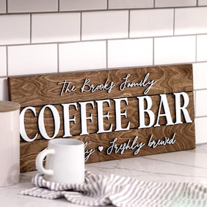Puede incluir: Un letrero de madera con el texto "The Brooks Family Coffee Bar" en letras blancas. El letrero tiene un acabado de madera marr&oacute;n r&uacute;stico y el texto "Daily Freshly Brewed" est&aacute; escrito en cursiva debajo del texto principal.