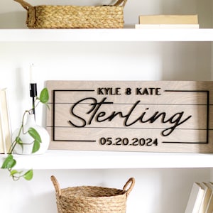 May include: A white bookshelf with two shelves. The top shelf has a woven basket and a book. The middle shelf has a wooden sign with the text "Kyle & Kate Sterling 05.20.2024" and a white vase with a green plant. The bottom shelf has a woven basket and three books.