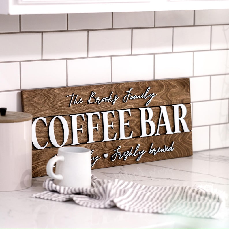 Puede incluir: Un letrero de madera con el texto "The Brooks Family Coffee Bar" en letras blancas. El letrero tiene un acabado de madera marr&oacute;n r&uacute;stico y el texto "Daily Freshly Brewed" est&aacute; escrito en cursiva debajo del texto principal.