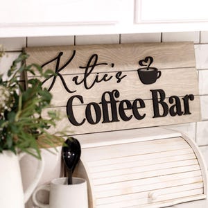 May include: A wooden sign with the words "Katie's Coffee Bar" in black lettering. The sign has a light brown wood grain background and a black coffee cup icon. A white mug, a white breadbox, and a white pitcher are in the foreground.
