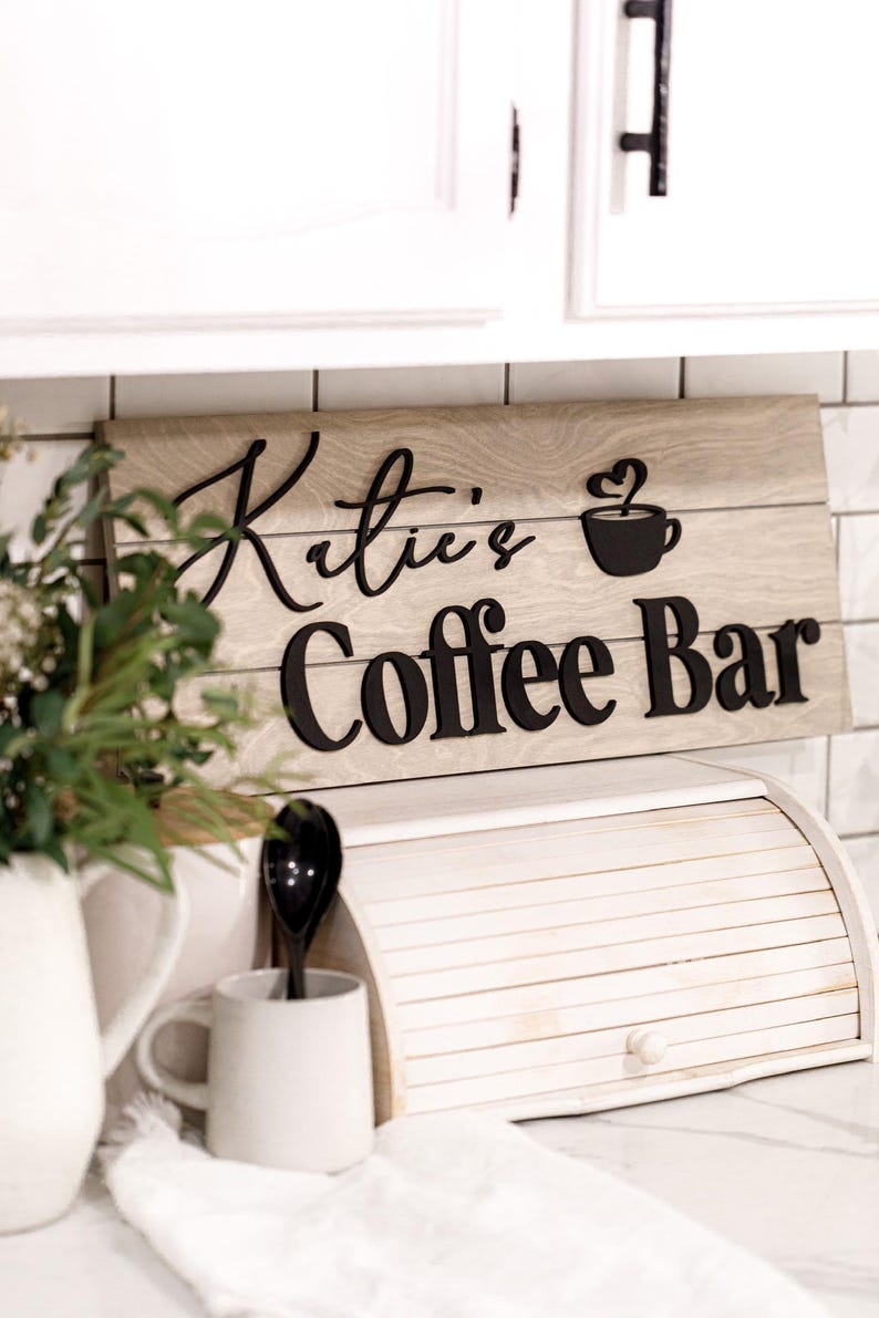May include: A wooden sign with the words "Katie's Coffee Bar" in black lettering. The sign has a light brown wood grain background and a black coffee cup icon. A white mug, a white breadbox, and a white pitcher are in the foreground.