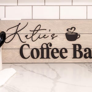 May include: Wooden sign with the words "Katie's Coffee Bar" in black lettering. The sign features a coffee cup with a heart-shaped steam design. The background is a light wood grain.