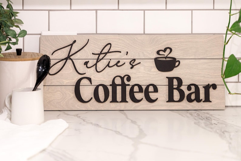 May include: Wooden sign with the words "Katie's Coffee Bar" in black lettering. The sign features a coffee cup with a heart-shaped steam design. The background is a light wood grain.