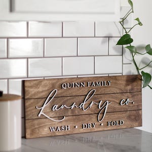 May include: A wooden sign with the words "Quinn Family Laundry Co." in white cursive lettering. The sign also has the words "Wash, Dry, Fold" in white lettering.