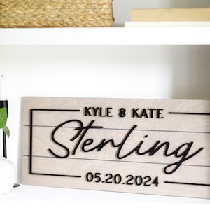 May include: A light gray wooden sign with black lettering. The sign reads "Kyle & Kate Sterling 05.20.2024".