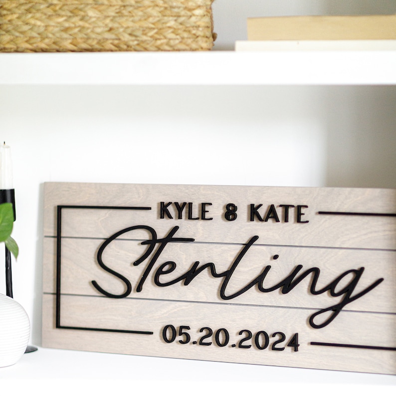 May include: A light gray wooden sign with black lettering. The sign reads "Kyle & Kate Sterling 05.20.2024".