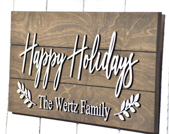 May include: A wooden sign with the text "Happy Holidays" in white script lettering. The sign also includes the text "The Wert Family" in white lettering. The sign is decorated with two white stylized sprigs of leaves.