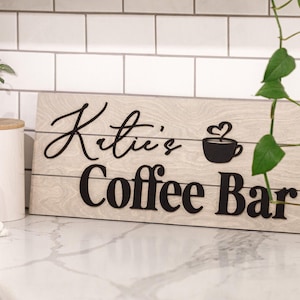 May include: Wooden sign with the text "Katie's Coffee Bar" in black lettering. The sign features a coffee cup with a heart-shaped steam design. The sign is light brown with a wood grain pattern.