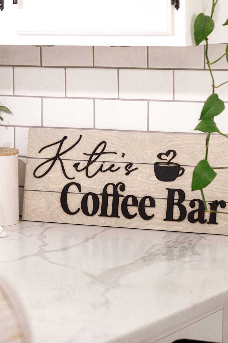 May include: Wooden sign with the text "Katie's Coffee Bar" in black lettering. The sign features a coffee cup with a heart-shaped steam design. The sign is light brown with a wood grain pattern.