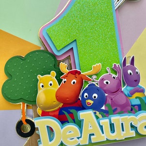 May include: A green glitter number one cake topper with a pink, blue, and gold border. The topper features characters from the children's show "The Backyardigans" including Uniqua, Tyrone, Pablo, Tasha, and Austin. The topper also includes a green tree with a gold swing and the name "DeAura" in yellow letters.