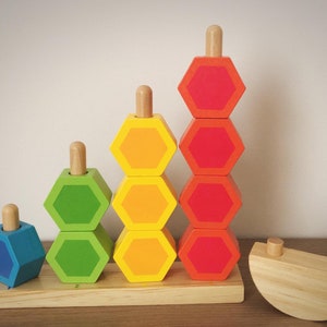 Può includere: Un giocattolo di impilamento in legno con blocchi esagonali blu, verdi, gialli e rossi. I blocchi sono impilati a forma di piramide su una base in legno. C'è un pezzo separato in legno a forma di semicerchio con due pomelli in legno.