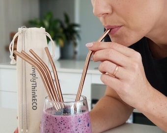 May include: Rose gold metal straws in a glass of purple smoothie. A canvas bag with the text "SimplyThinkECO" is next to the glass. Several blueberries are on the white countertop. One straw is being used to drink the smoothie.