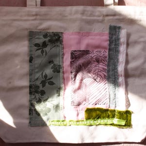 Pu&ograve; includere: Una borsa tote in tela bianca con un design a patchwork. Il design presenta una stampa floreale, un tessuto rosa con una grafica in bianco e nero e un nastro di velluto verde.