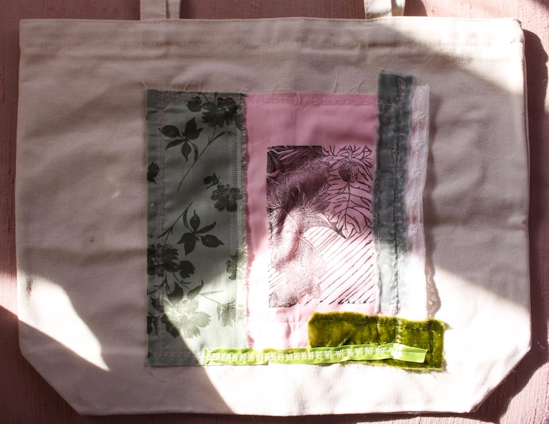 Pu&ograve; includere: Una borsa tote in tela bianca con un design a patchwork. Il design presenta una stampa floreale, un tessuto rosa con una grafica in bianco e nero e un nastro di velluto verde.