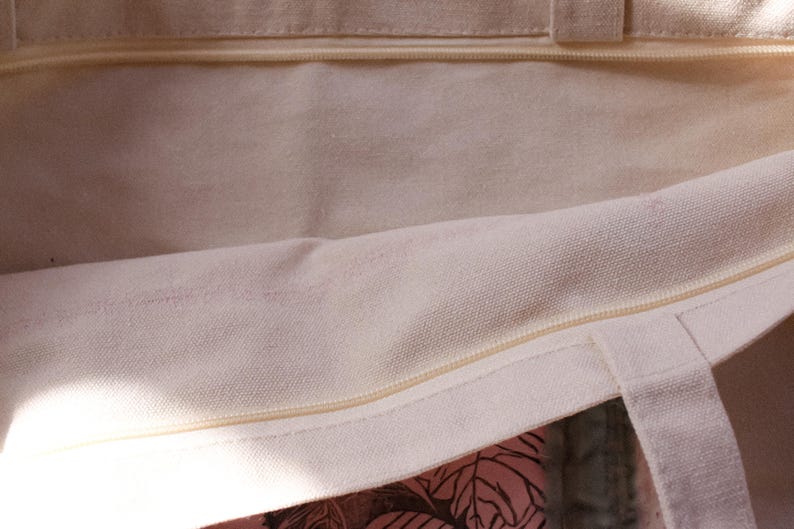 Pu&ograve; includere: Primo piano dell'interno di una borsa tote in tela bianca con chiusura a zip. La borsa non &egrave; foderata e ha un aspetto naturale non sbiancato.